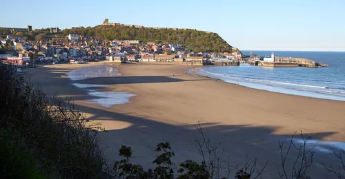 Scarborough beach evening This landscape photograph captures Scarborough beach in the evening during spring, as evidenced by the lush greenery and the long shadows cast by the setting sun. The expansive sandy shoreline leads to the waters of the North Sea, with gentle waves approaching the coast. Above the town, Scarborough Castle stands prominently atop a hill, offering a historic landmark that overlooks the picturesque coastal scene. The town of Scarborough, located in Yorkshire, features a collection of traditional buildings clustered between the beach and the castle, creating a harmonious blend of coastal and urban elements. The peaceful ambiance and soft light of early evening give the beach a calm, inviting atmosphere, characteristic of Yorkshire’s scenic beauty.